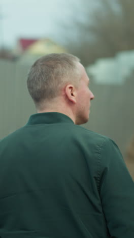 a close view of a man walking along a fence, captured from behind, with a view of back of his head and jacket, with a blurred background of an industrial area