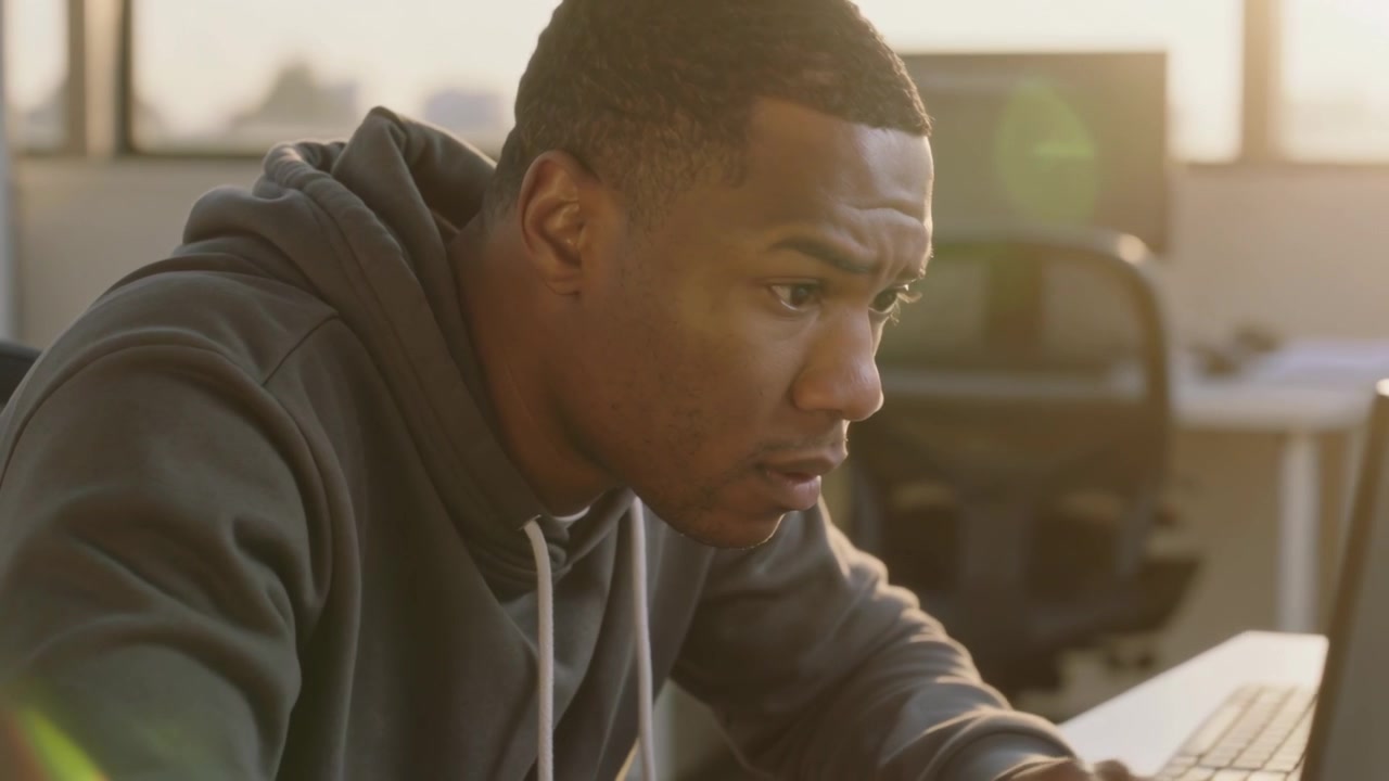 Reading man in dark gray hoodie leaning toward laptop at office desk, touching keyboard, responding