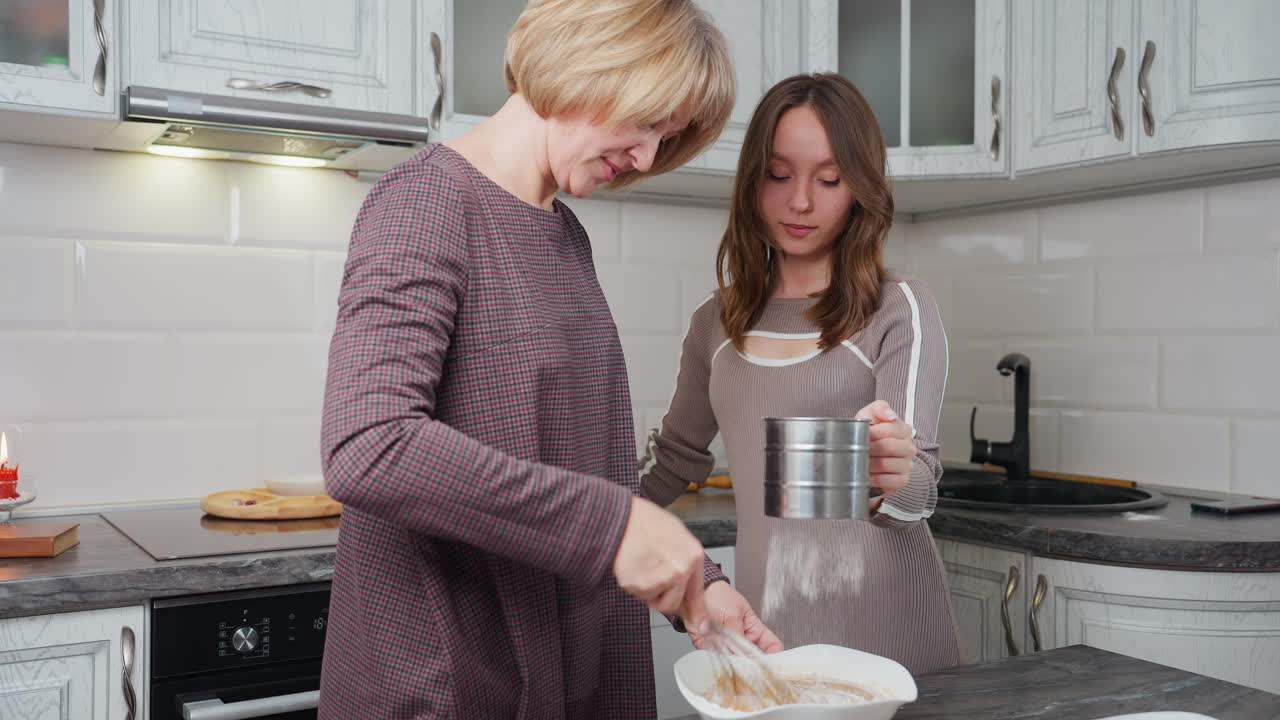 Young girl helps her mother in kitchen, sifting flour into mixing bowl while mother stirs ingredients, warm family moment in bright, modern kitchen showcasing teamwork