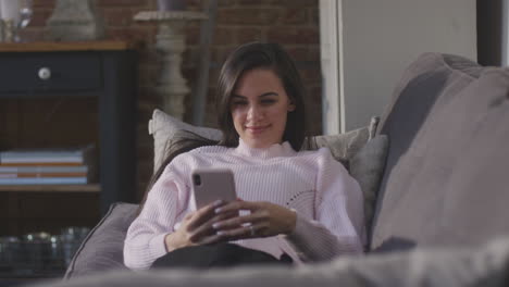 mujer sonriente relajándose en el sofá en casa revisando las redes sociales en el teléfono móvil