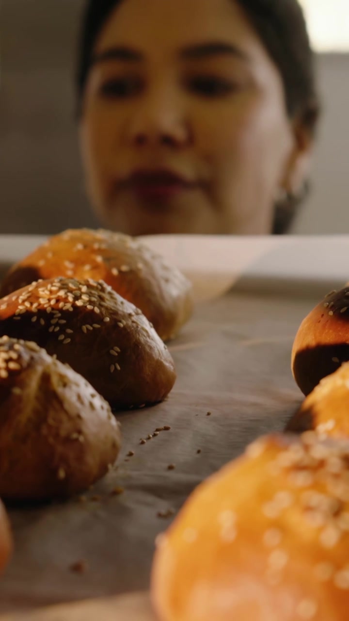 Vertical video: Hand entering cueing chef in dark top checking sesame rolls done at bench with pan