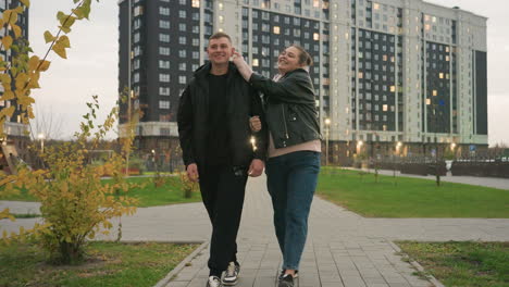 Two lovers walking on paved path as wife holds husband arm and gently tickles his ear causing both to laugh warmly surrounded by residential buildings street lights and autumn trees