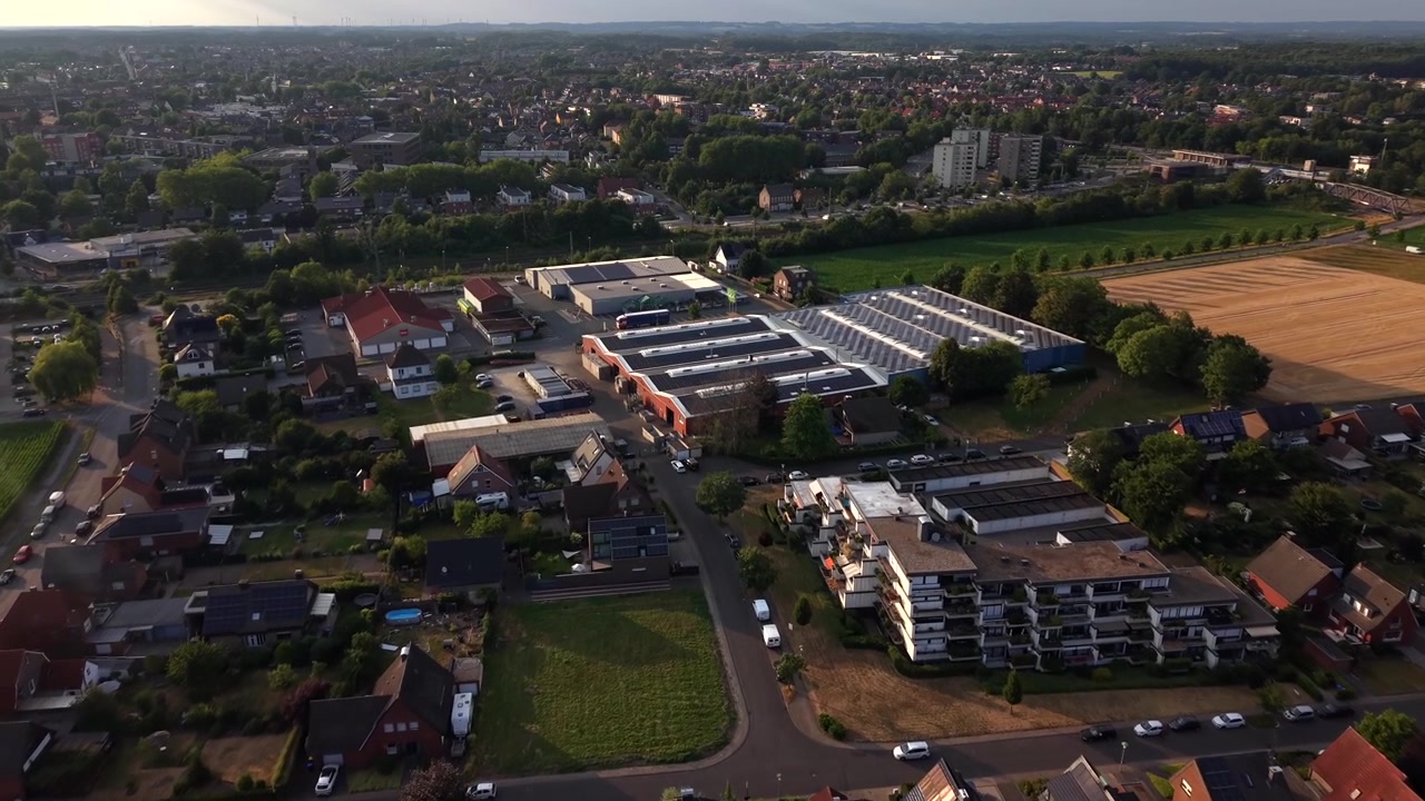 Zonnepanelen bedekken rijen daken van woonhuizen in een klein Duits stadje, bedrijventerrein en open landbouwgrond verspreid langs rustige buitenwijken tijdens een warme gouden zonsondergang. Brede luchtopname met drone