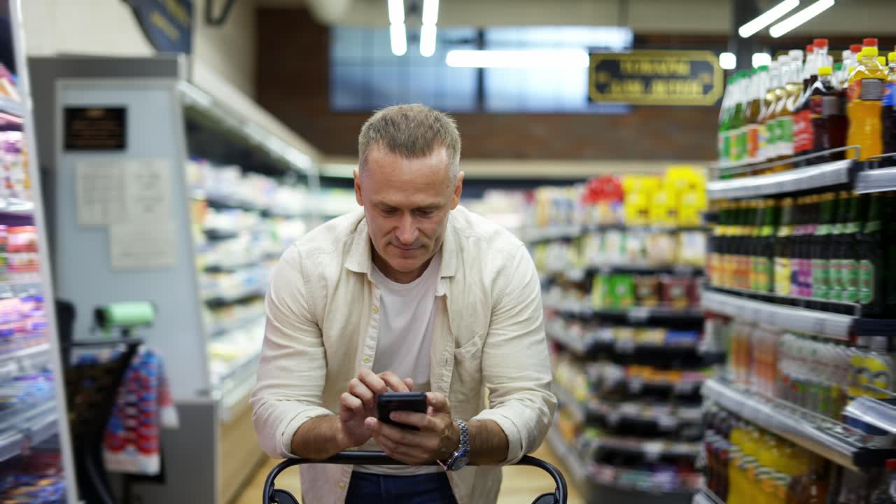 Un uomo di mezza età cammina per il supermercato, porta un carrello e tiene un telefono in mano. Andare al supermercato. Quotidiano.
