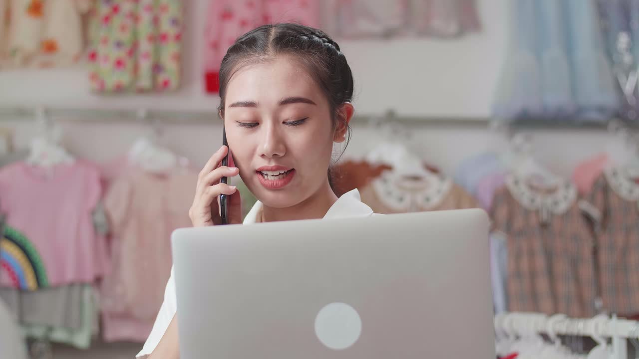 아동 의류 가게의 아시아 여성 소유자가 노트북을 사용하여 스을 확인하는 동안 스마트폰으로 이야기하고 있습니다.