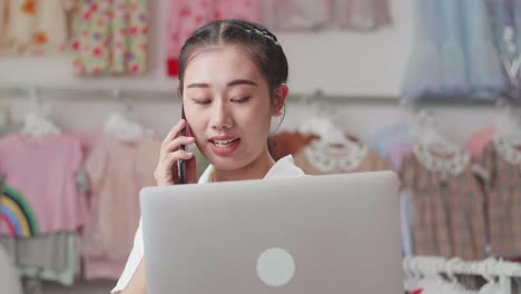 un primer plano de una mujer asiática dueña de una tienda de ropa para niños hablando en su teléfono inteligente mientras usa una computadora portátil para comprobar las existencias en los rieles