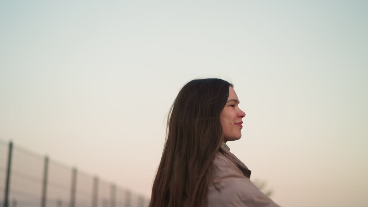 una vista de cerca de una mujer joven con una chaqueta de melocotón, disfrutando de un momento de patinaje con una expresión alegre en su rostro. el fondo presenta un cielo suave y pastel al atardecer