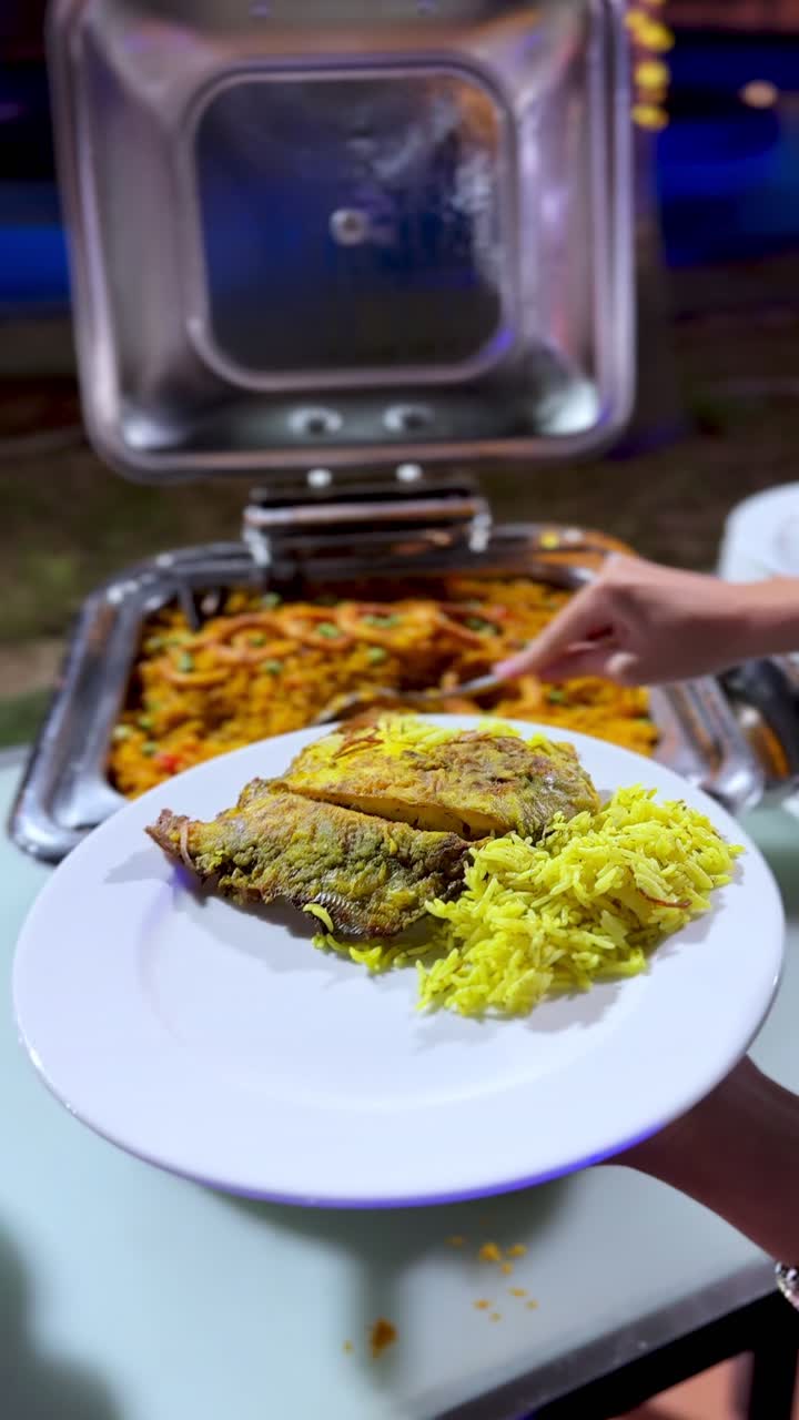 Close-up of a guest serving seafood paella with calamari rings from a silver chafing dish. Hot rice meal at an outdoor evening resort buffet with blue ambient lighting