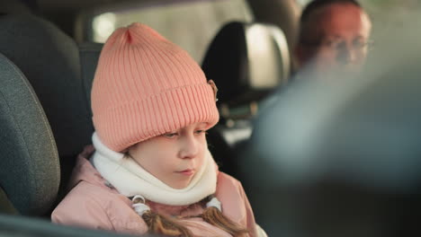 a close-up shot of a young girl dressed warmly in a pink beanie and jacket, sitting quietly in a car. the image captures her calm and focused expression, with a man subtly visible in the background