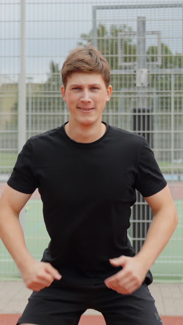 Young man engaging in a squat exercise outdoors, smiling and enjoying a physical workout on a sports ground, focusing on fitness, health, and well being, vertical camera track, slow motion shot