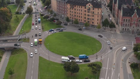 Drone-footage-of-traffic-driving-in-green-grass-roundabout-in-Roslagstull,-Stockholm,-Sweden-on-overcast-day-with-lush-green-grass-and-trees-at-start-of-fall-revealing-city-skyline