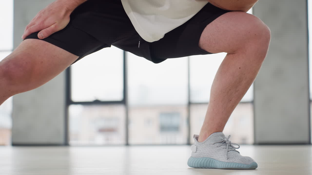 Waist view of fitness trainer performing side lunge warm up exercise in bright modern gym wearing white tank and black shorts balancing on light wooden floor beneath hanging straps and large windows