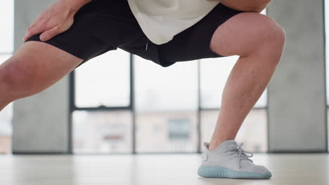 waist view of fitness trainer performing side lunge warm up exercise in bright modern gym wearing white tank and black shorts balancing on light wooden floor beneath hanging straps and large windows