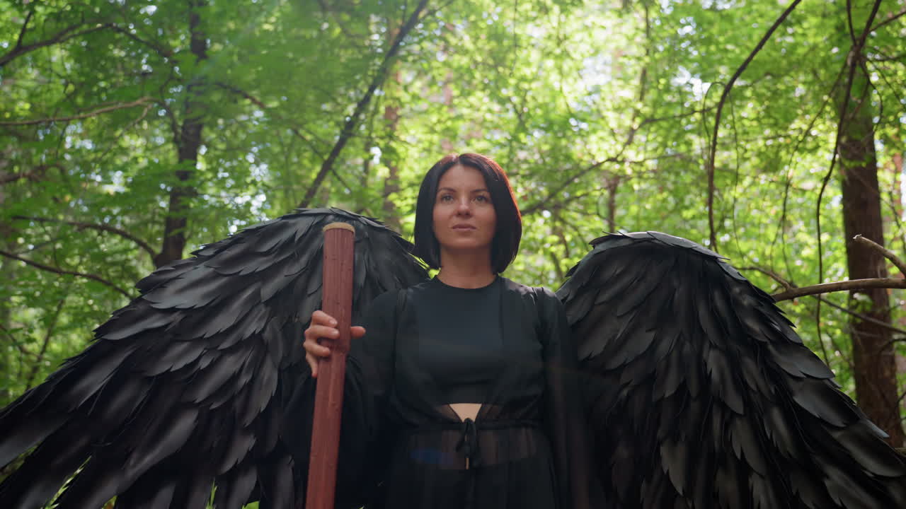 Confident woman in black bird costume walks through forest holding wooden staff, surrounded by glowing sunlight and green leaves, creating mysterious cinematic atmosphere of strength