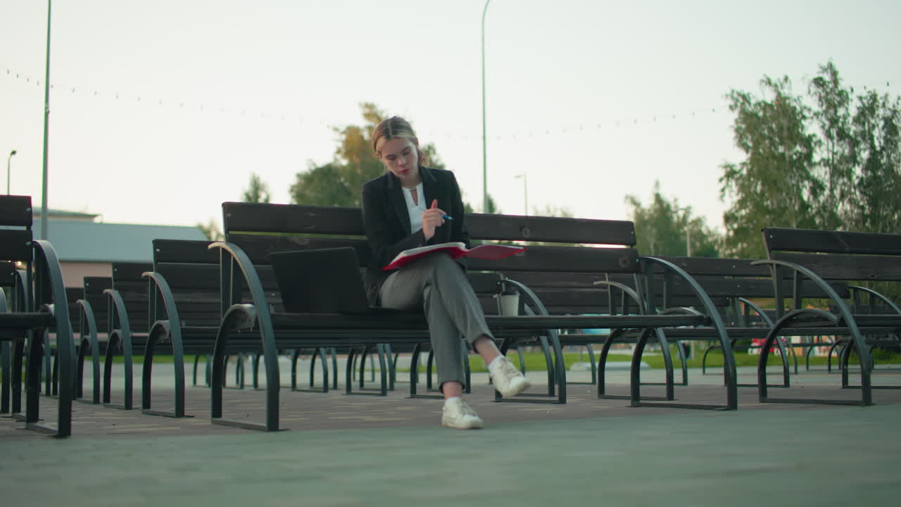 Student seated alone on outdoor bench deeply focused reading large open book while holding pen with laptop and coffee cup by her side in peaceful urban area surrounded by empty benches