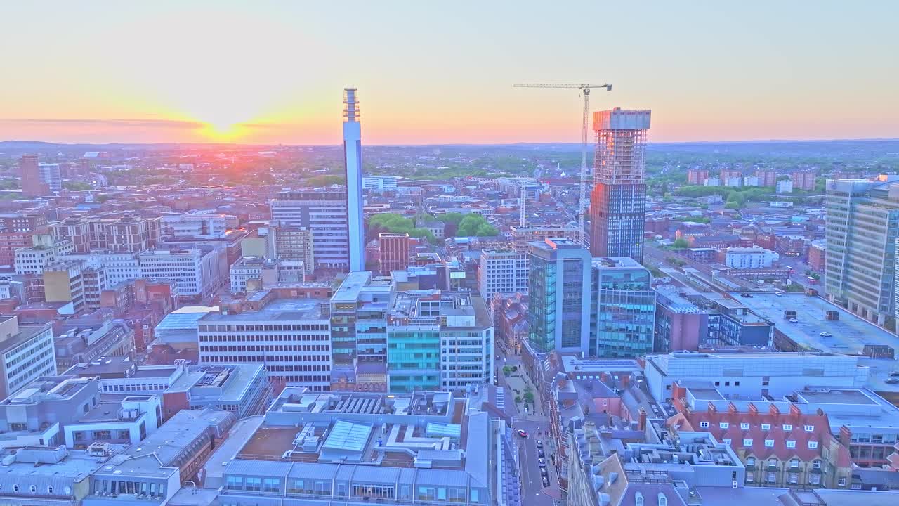 Drone footage captures Birmingham’s cityscape at golden hour, highlighting the BT Tower, modern office buildings, and skyscrapers under construction as the sun sets over the urban landscape