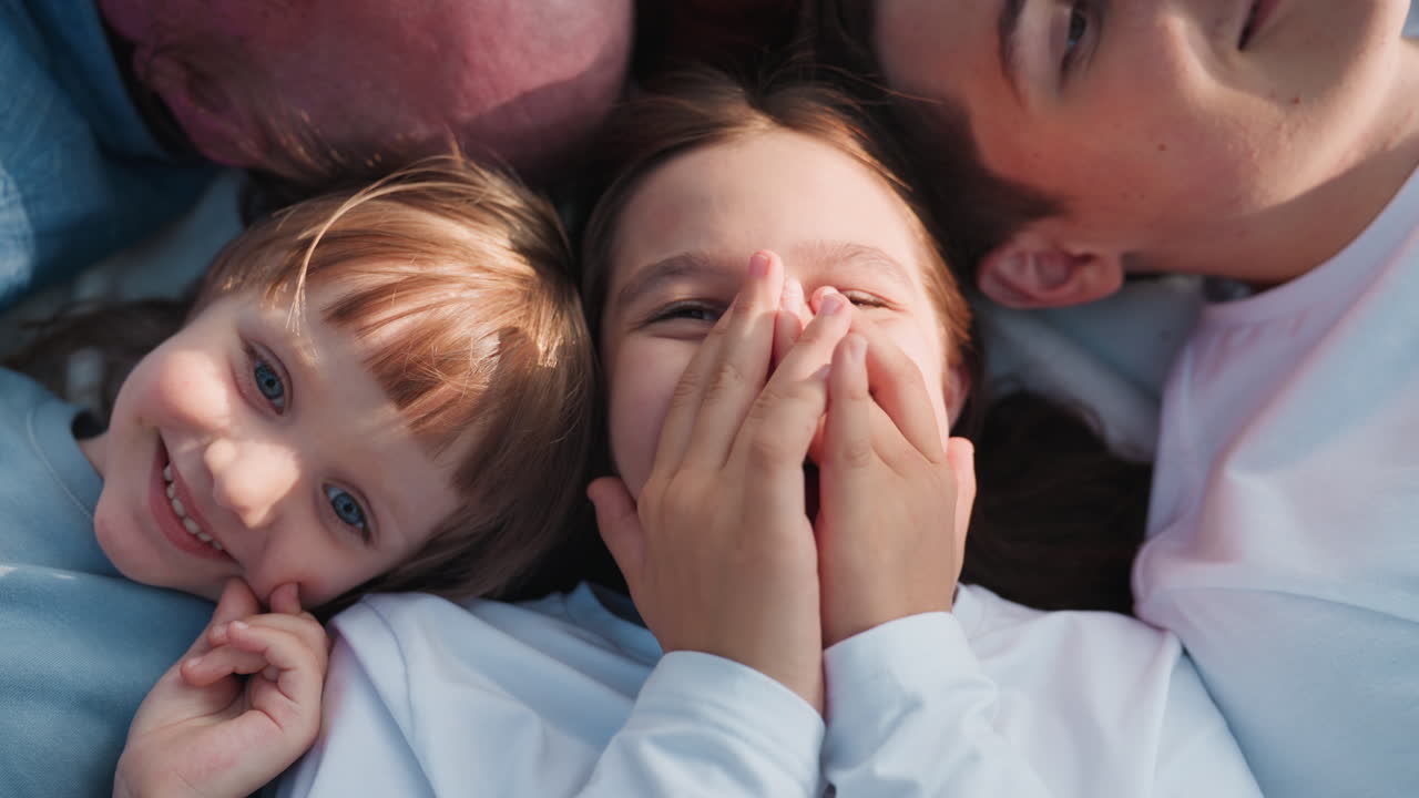 Luftfoto af en teenagepige, der ligger på ryggen mellem bror og søster og ler glad, mens de laver sjov sammen og udviser varme, humor, familiebånd og let hjerte.