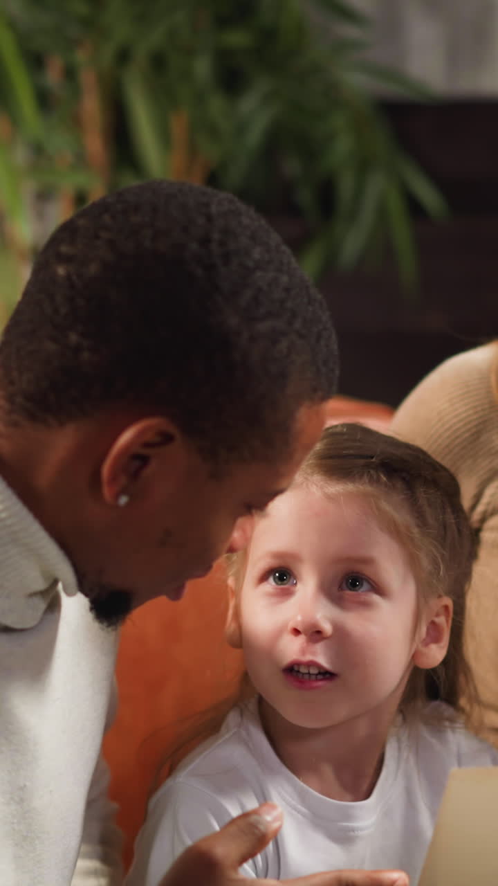 Happy blonde woman turns page of photo album and black father shows picture of grandfather to preschooler girl in living room closeup slow motion