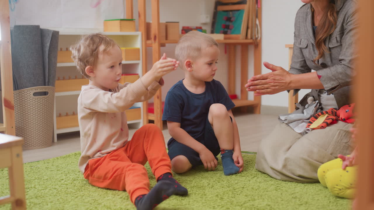 Preschool scene with woman guiding toddlers through hand movement routine, one child imitates raised arms while boy in blue watches, seated on green rug in classroom