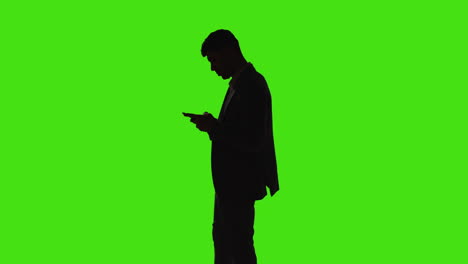 silhouette of businessman sending or reading text message or scrolling online on mobile phone standing against green screen with low key lighting