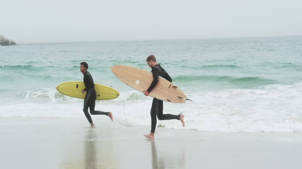 vista lateral de dos surfistas masculinos corriendo con una tabla de surf en la playa 4k