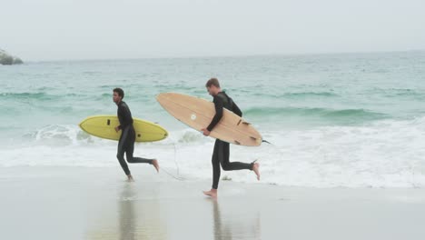 Vista-Lateral-De-Dos-Surfistas-Masculinos-Corriendo-Con-Tabla-De-Surf-En-La-Playa-4k