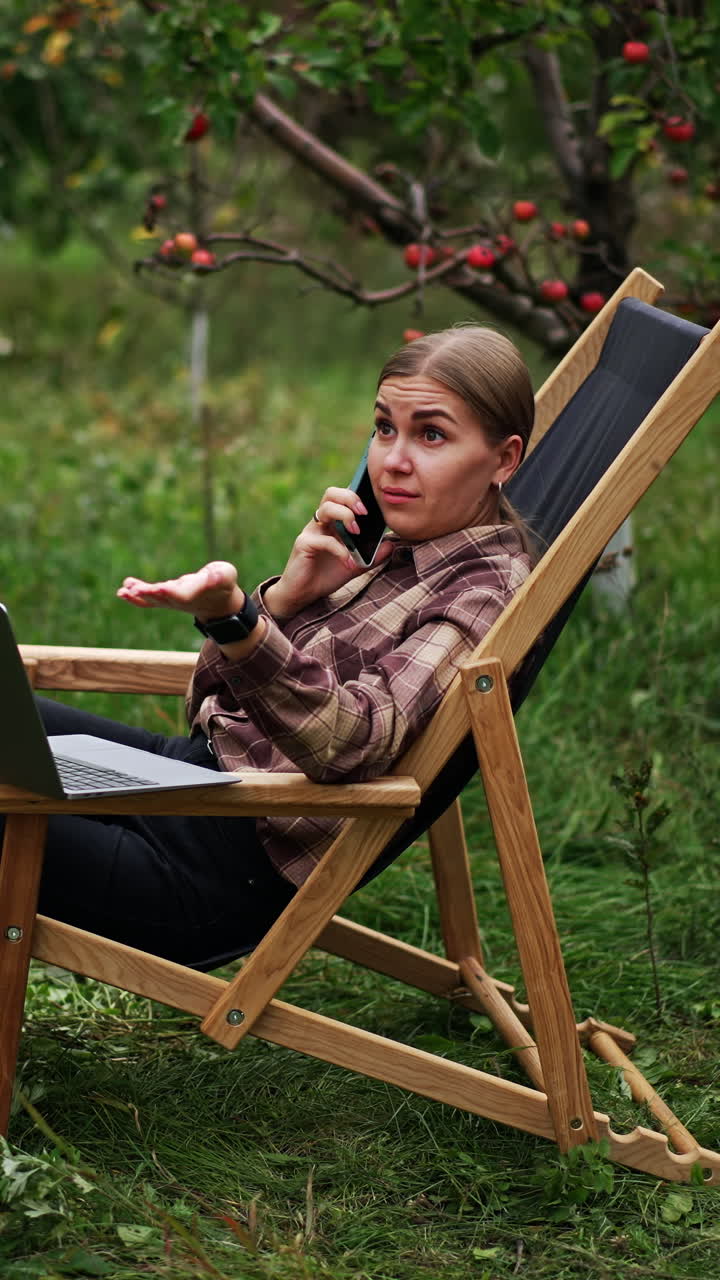 Busy lady working freelance speaks emotionally on the phone. Lady is puzzled about conversation. Opened laptop in front of woman. Vertical video