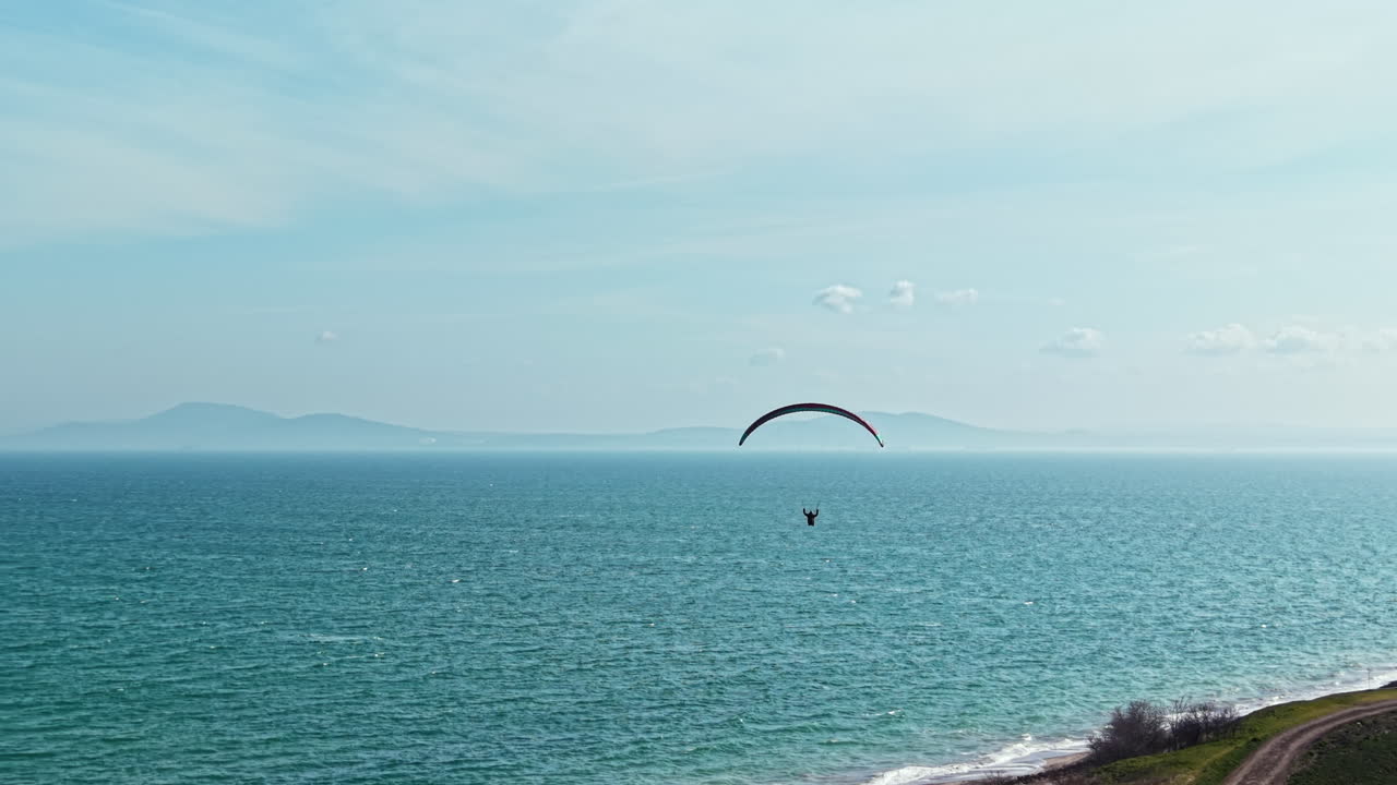 Paraglider flyr over det rolige blå havet nær kystlinjen med fjerne fjell under en klar himmel på ettermiddagen.