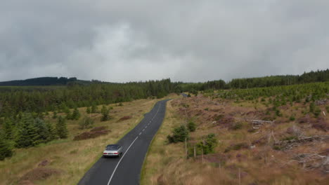 Following-silver-vintage-sports-car-driving-on-curvy-road-in-wooded-landscape.-Overcast-sky-on-rainy-day.-Ireland