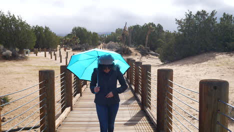 pretty woman walking across a bridge with a blue rain umbrella with stormy, cloudy skies above slow motion