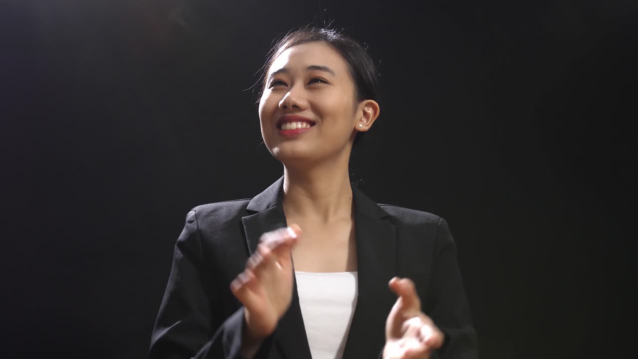 블랙 스크린 스튜디오에 서서 손을 박고 주변을 둘러보는 미소 짓는 아시아 스피커 여성