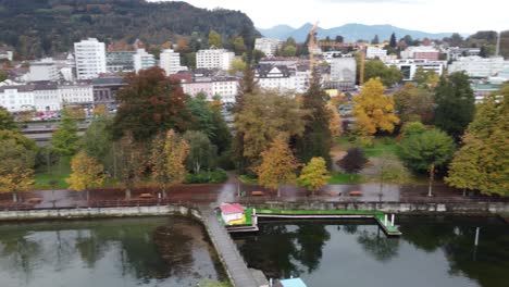 Picturesque-city-of-Bregenz-with-its-typical-alpine-houses-surrounded-by-trees-and-on-the-shore-of-the-lake-its-small-docks-with-its-sky-overflown-with-birds,-aerial-view