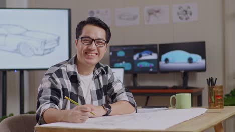 side view of asian male smiling to the camera while working on a car design sketch on table in the studio with tv and computers display 3d electric car model