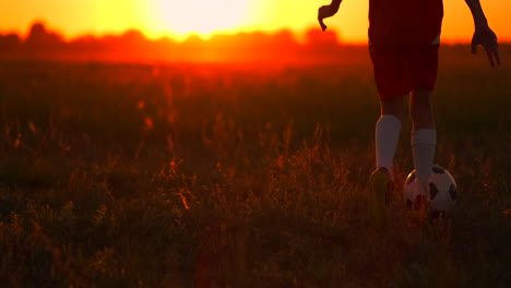 un jeune joueur de football dans un t-shirt rouge et un short qui court avec le ballon au coucher du soleil sur le terrain sur l'herbe. le concept de jeunes joueurs. le jeune joueur de foot rêve d'une carrière professionnelle et s'entraîne sur le terrain.