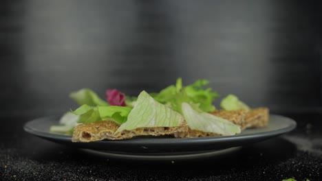 Hojas-De-Ensalada-Verde-Cayendo-Sobre-Pan-Tostado,-Primer-Plano-De-Fondo-Gris