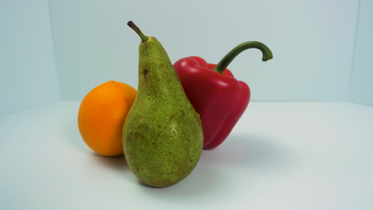 Frisk stor grønn pære, rød søt paprika, saftig appelsin roterer sakte på en lys blå bakgrunn, sunt matkonsept, nærbilde, kameraet roterer til venstre.
