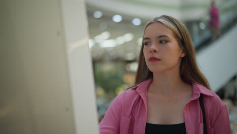 lady in pink dress with black handbag intensely focused on using screen in well-lit mall, the background features blurred lights and escalators with people, as she appears focus on the task