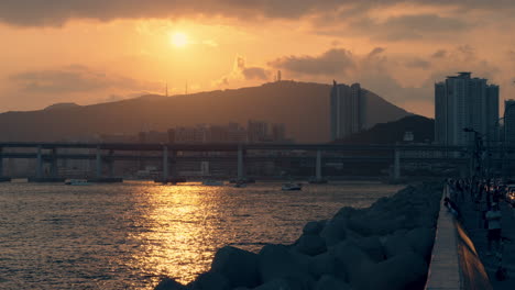 sunset at busan marina bay with a view of gwangan bridge or gwangandaegyo, tourists admire setting over the mountain sun seafront promenade, travel yachts cruising through reflected in sea sunlight
