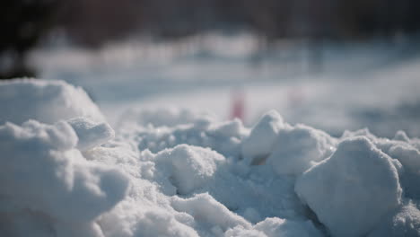 closeup of powdery snow clumps shimmering in sun with blurred figures walking in background on winter path, delicate crystalline texture and soft bokeh hints of human presence in cold landscape