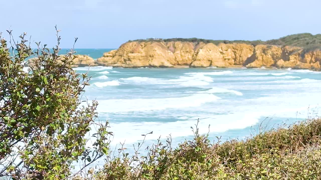 Waves crash against cliffs in Torquay, Australia. Bright daylight illuminates the coastal scenery, captured with steady camera movement