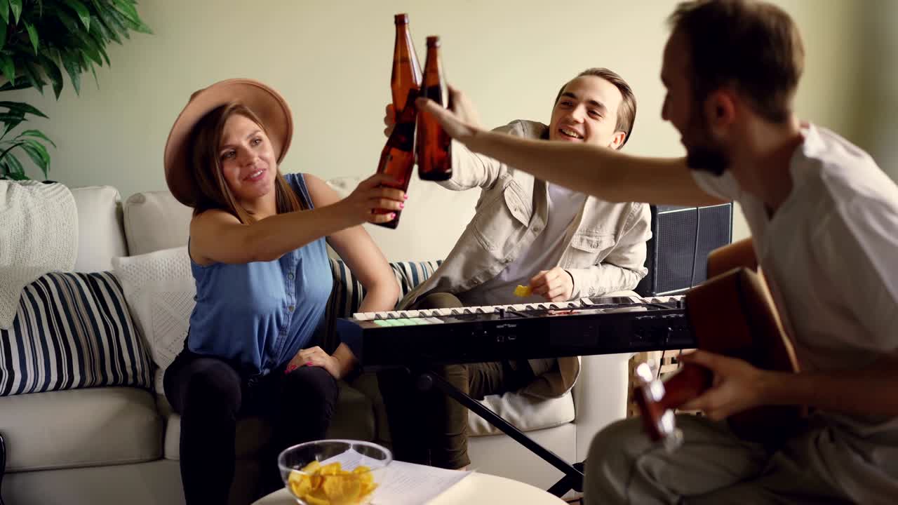 성공한 뮤지션들은 새로운 앨범을 축하하고, 병을 걱거리며, 홈 스튜디오에서 이야기하고 웃고 있습니다. 전기 악기, 간식, 음료가 보입니다.