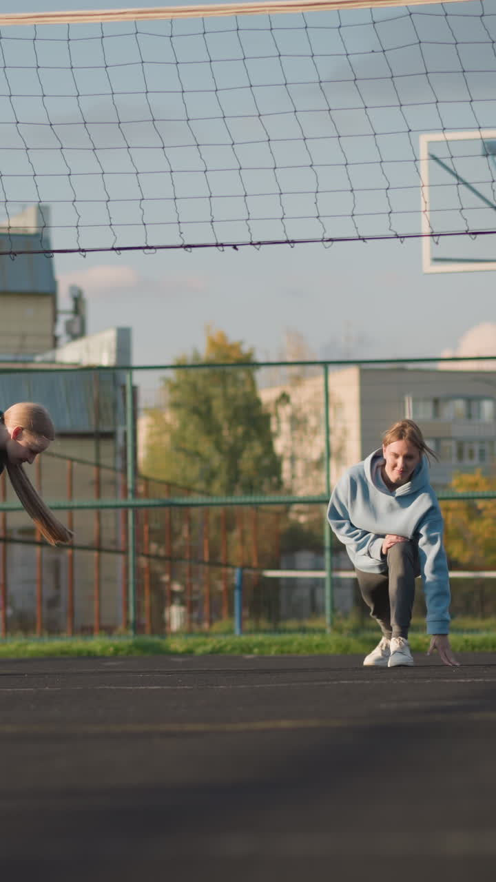 primer plano de voleibol en el suelo con fondo borroso de atletas estirándose y haciendo ejercicio al aire libre, red de voleobol y cancha deportiva visibles con edificios distantes bajo un cielo despejado