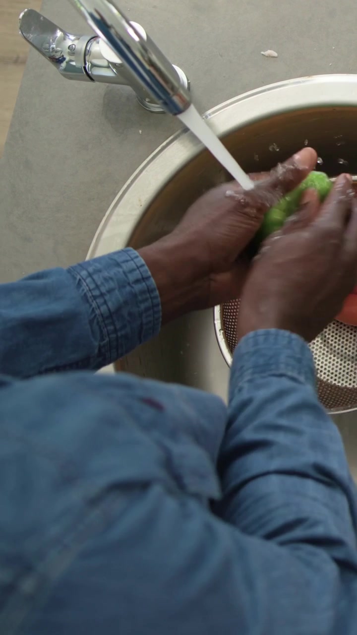 Video verticale: Uomo in jeans che sciacqua carote e peperoni sotto l'acqua corrente in uno scolapasta per la preparazione.