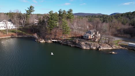 Lakeside-colonial-homes-on-the-water,-cars-cruising-on-a-road-with-blue-sky-and-calm-waters-among-evergreen-forests-and-rolling-hills-and-a-bird-flying-by