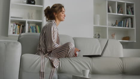 girl making video call on living room couch. happy woman waving hand at laptop