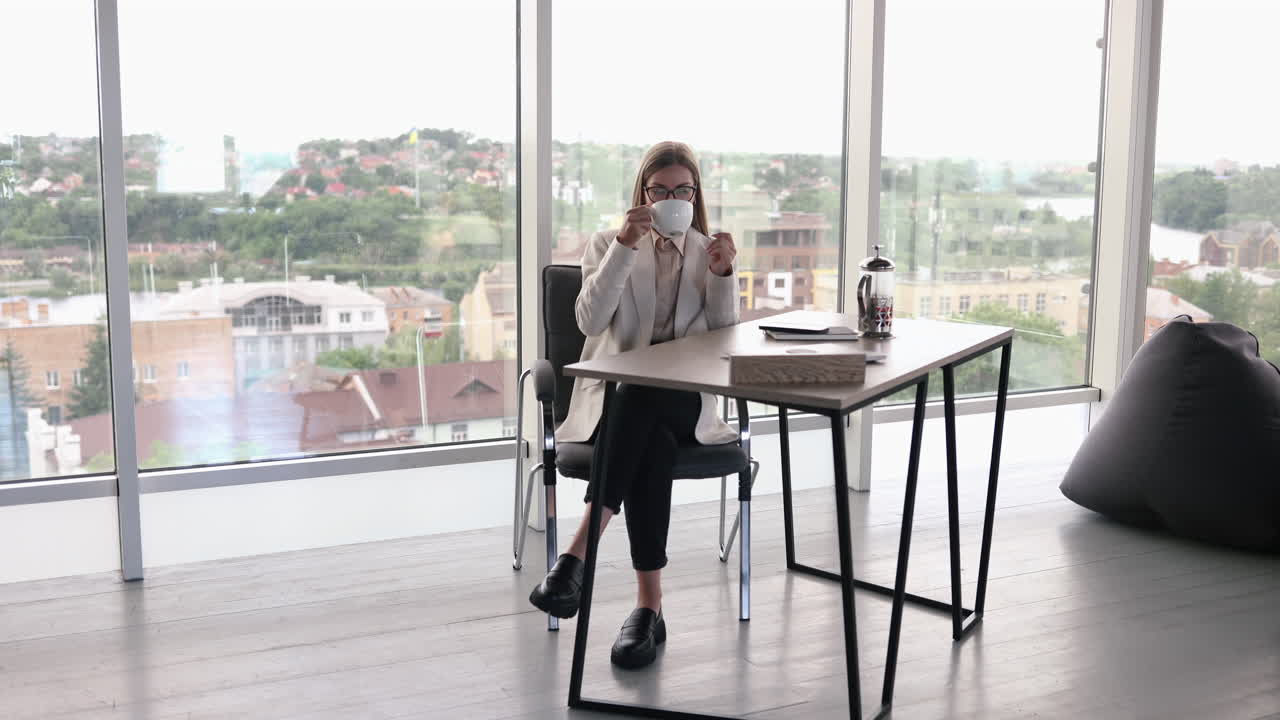 Bella giovane donna con gli occhiali che lavora in un ufficio luminoso e beve tè al laptop. Affascinante signora dai capelli lunghi con gli occhiali che parla al telefono
