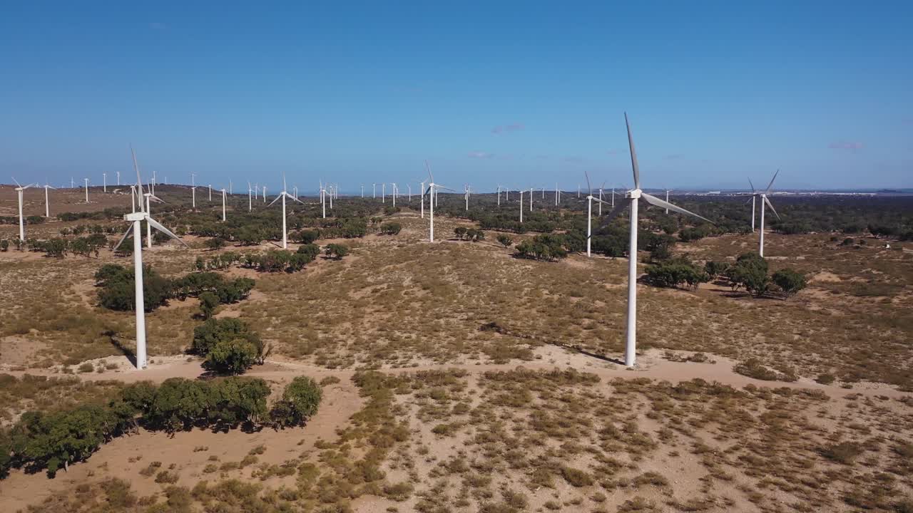Imagem aérea de drone de um parque eólico com várias turbinas eólicas brancas girando e gerando eletricidade limpa, situado em uma paisagem árida e seca com vegetação esparsa sob um céu azul claro, Marrocos