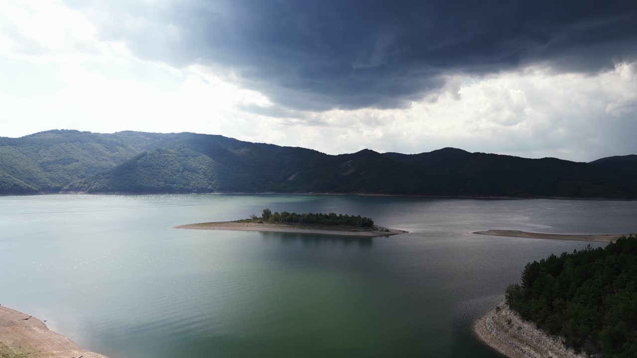 저수지에 있는 섬의 극적인 공중 촬영, 폭풍우 구름이 머리 위에 모여 있고 배경에는 산이 있습니다.