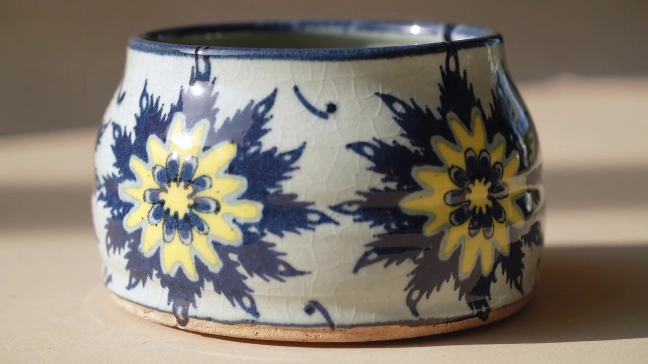 La telecamera si sposta, facendo ruotare un vaso di ceramica su un tavolo beige, che mostra motivi blu e gialli.