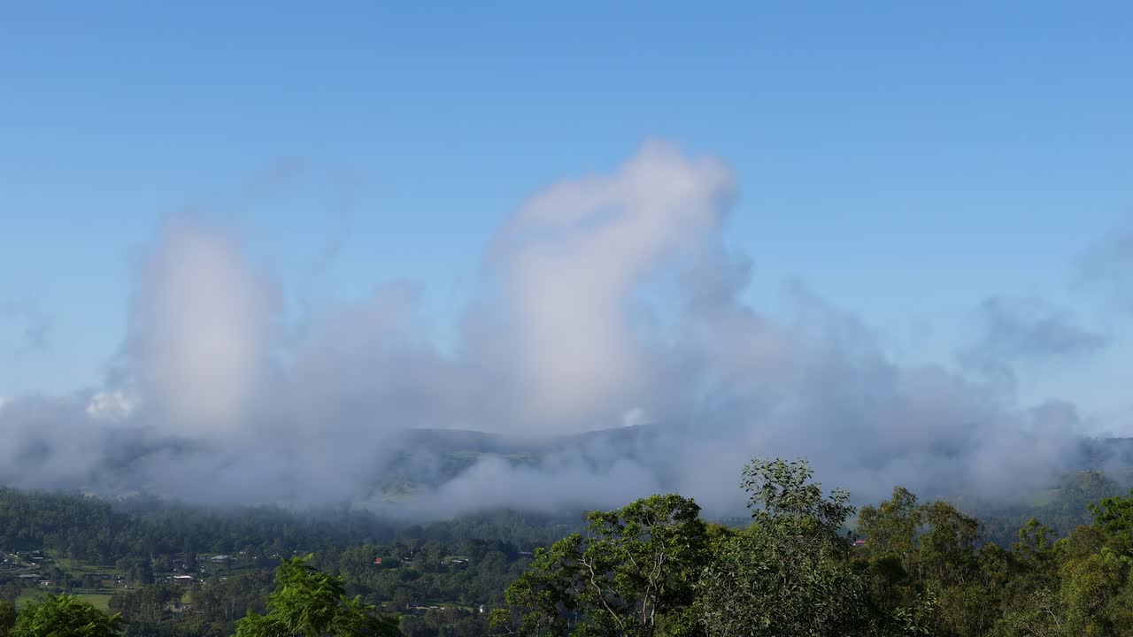 Watch clouds gently move across a landscape of green hills and dense treetops under a clear blue sky.